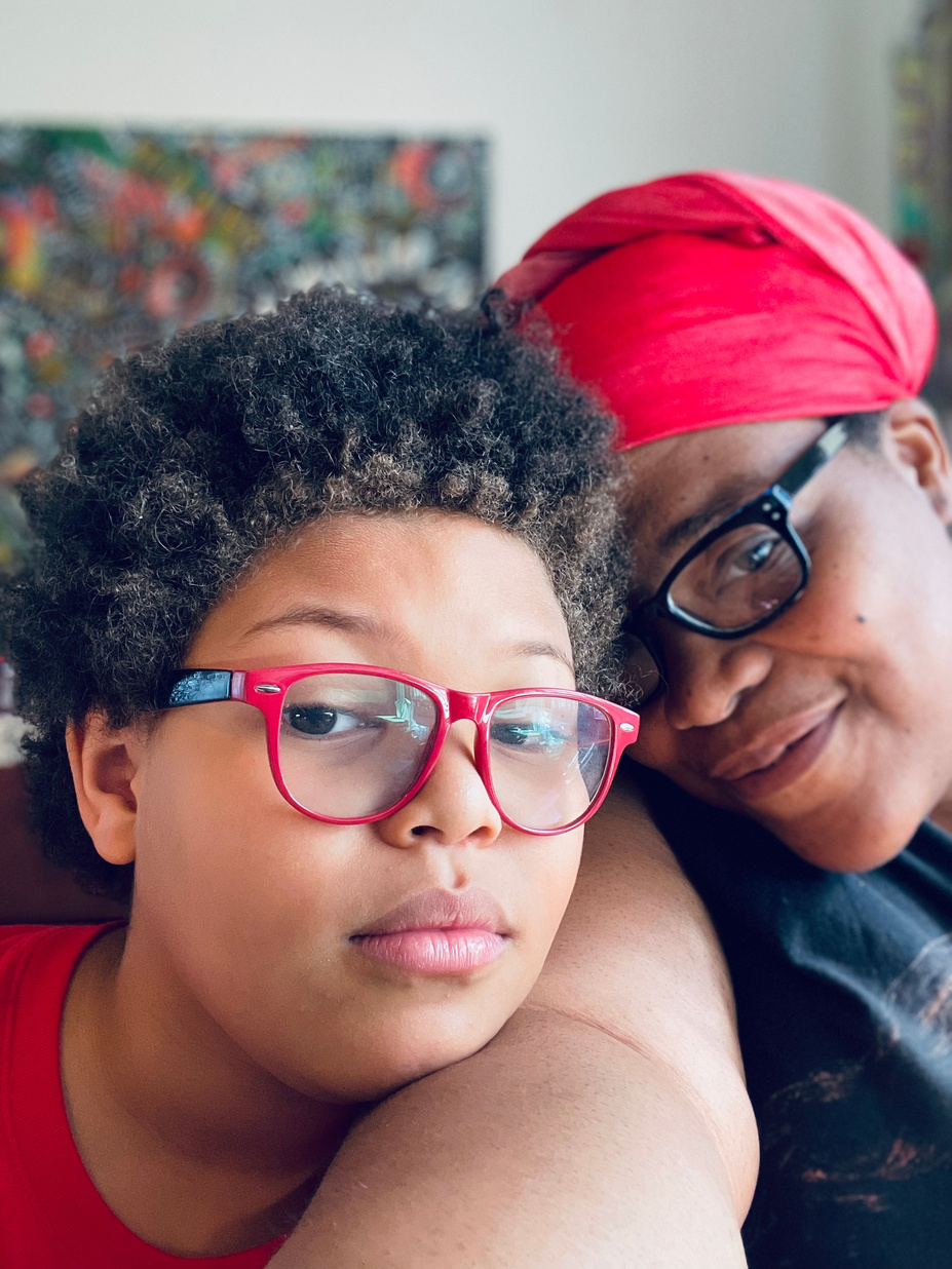 Bellamy and oldest child take a selfie looking contemplative. Child has on bright red glasses and has an afro, Bellamy have on black glasses and a bright red head wrap. There is abstract art behind them.