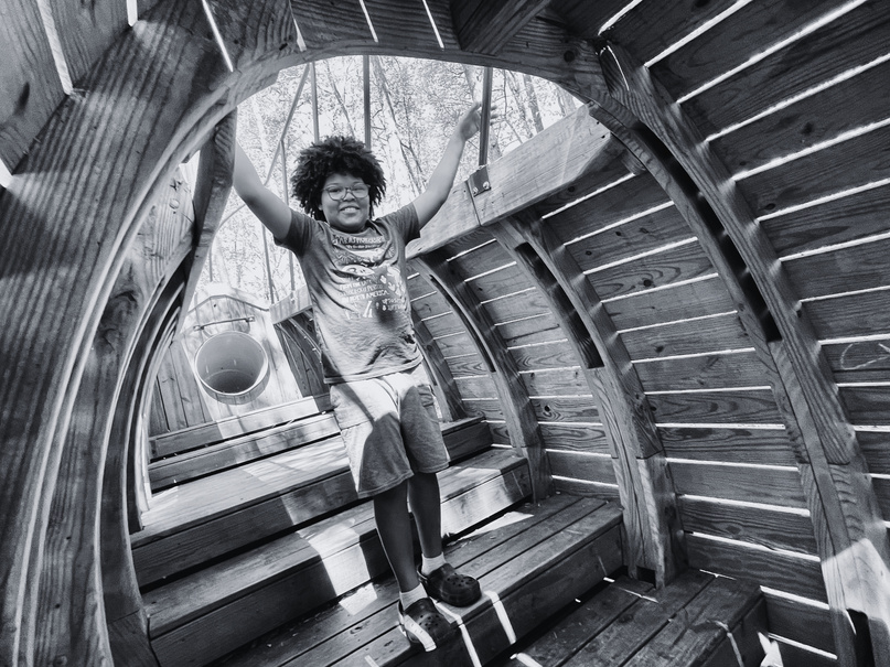Bellamy's child smiling inside a giant wooden fish at a playground. 