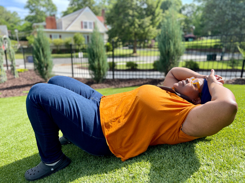 Bellamy Shoffner Black femme lays on turf looking up at the sky in mustard orange shirt and blue jeans. 